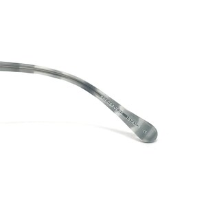 Puede incluir: Primer plano de la patilla de unas gafas. La patilla es de color gris claro con un patr&oacute;n veteado. El texto "SPECSAVERS" y "157431-1" est&aacute;n impresos en la patilla.