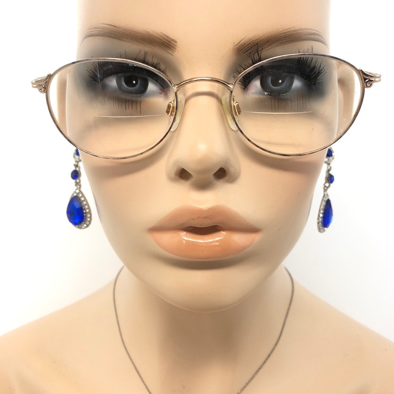 K&ouml;nnte beinhalten: Ovale goldfarbene Brille mit klaren Gl&auml;sern. Die Brille befindet sich auf einer Schaufensterpuppe. H&auml;ngende blaue Tr&auml;nenohrringe mit silbernen Akzenten sind ebenfalls zu sehen.