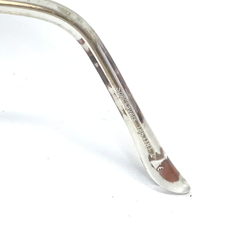 May include: Close-up of the arm of a pair of clear glasses with a gold trim. The arm is inscribed with "Specsavers 18531811".