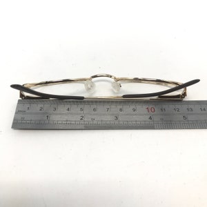 Pu&ograve; includere: Occhiali con montatura dorata e aste nere, mostrati con un righello per la scala. Gli occhiali hanno naselli trasparenti e sono lunghi circa 13 cm.