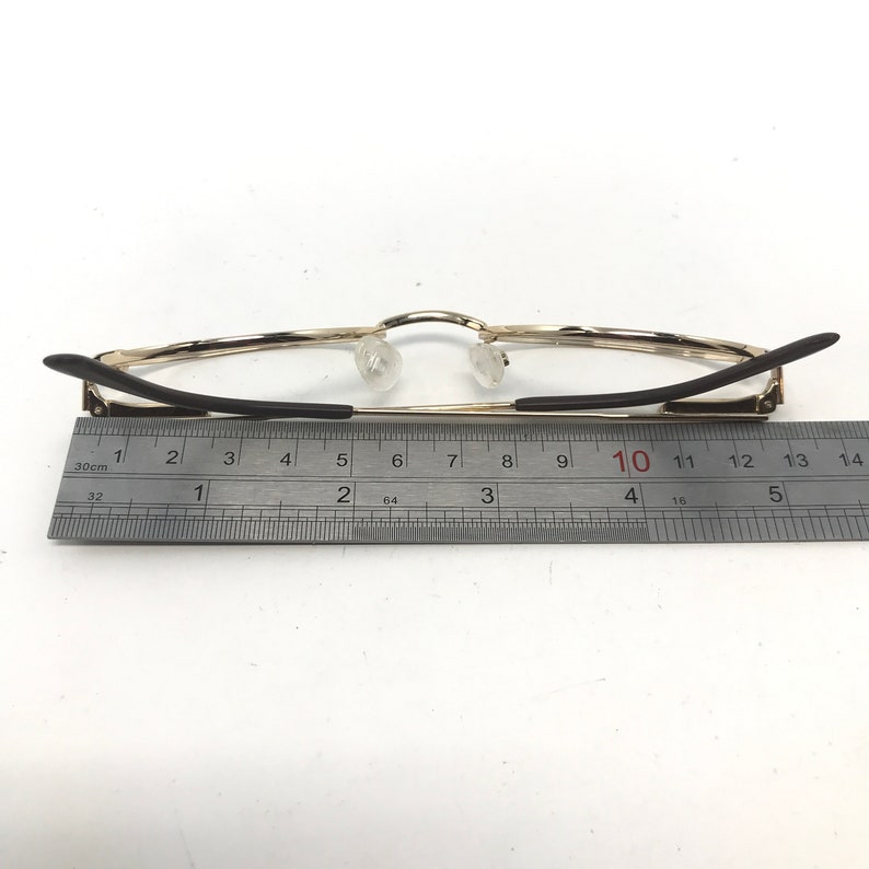 Pu&ograve; includere: Occhiali con montatura dorata e aste nere, mostrati con un righello per la scala. Gli occhiali hanno naselli trasparenti e sono lunghi circa 13 cm.