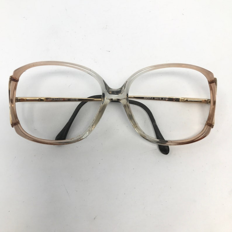 May include: Oversized eyeglasses with a gradient frame, transitioning from a light brown to a clear hue. The frames have gold-colored metal accents and black temples. The text on the frame reads "SPEC SAVERS" and "AB6051 2 54/15 A160".