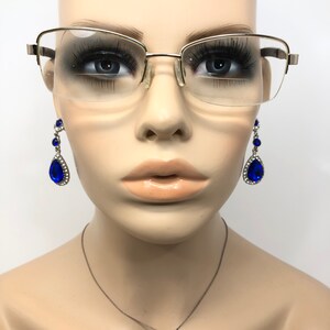 Pu&ograve; includere: Un busto di manichino con occhiali con montatura dorata e orecchini pendenti blu a goccia. Gli orecchini hanno una grande pietra blu circondata da pietre chiare pi&ugrave; piccole. &Egrave; visibile anche una collana d'argento.