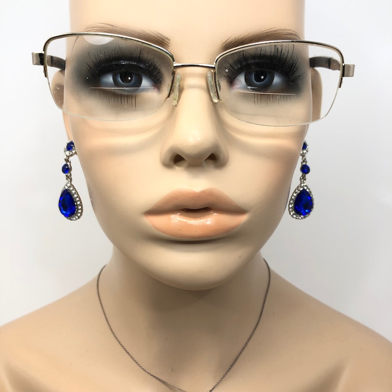 Pu&ograve; includere: Un busto di manichino con occhiali con montatura dorata e orecchini pendenti blu a goccia. Gli orecchini hanno una grande pietra blu circondata da pietre chiare pi&ugrave; piccole. &Egrave; visibile anche una collana d'argento.