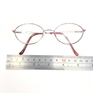 K&ouml;nnte beinhalten: Ovale Brille mit einem ros&eacute;goldenen Rahmen und braunen B&uuml;geln. Die Brille liegt auf einer wei&szlig;en Oberfl&auml;che neben einem Lineal. Der Rahmen hat ein filigranes, minimalistisches Design. Das Lineal zeigt Messungen in Zentimetern an.