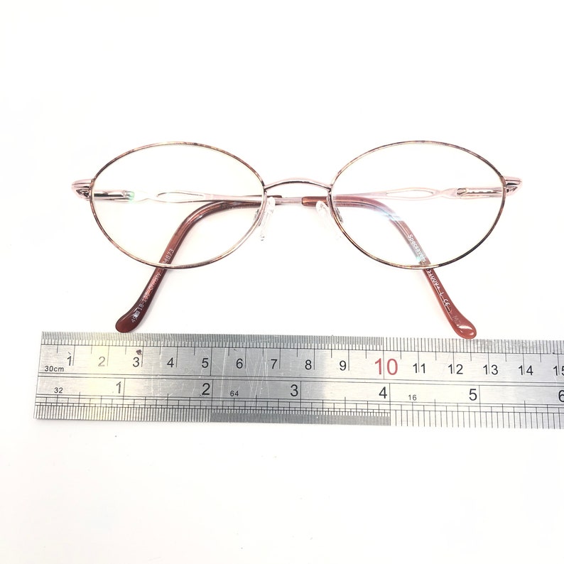 K&ouml;nnte beinhalten: Ovale Brille mit einem ros&eacute;goldenen Rahmen und braunen B&uuml;geln. Die Brille liegt auf einer wei&szlig;en Oberfl&auml;che neben einem Lineal. Der Rahmen hat ein filigranes, minimalistisches Design. Das Lineal zeigt Messungen in Zentimetern an.