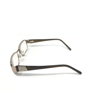 May include: A pair of eyeglasses with a rectangular frame. The frame is a metallic silver color with a brown arm. The arm has a decorative design with small, clear embellishments. The lenses are clear.