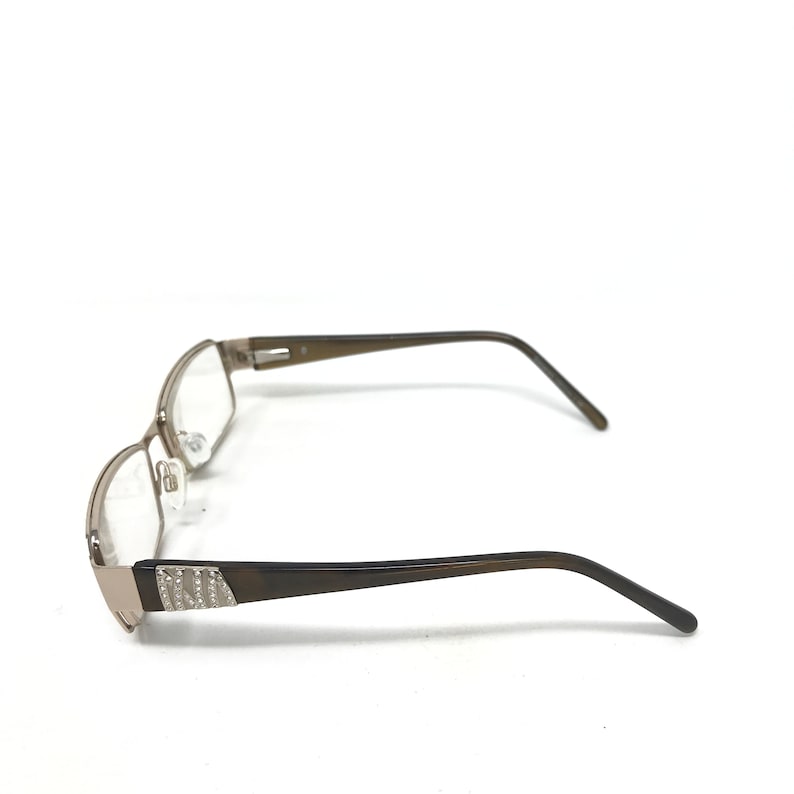 May include: A pair of eyeglasses with a rectangular frame. The frame is a metallic silver color with a brown arm. The arm has a decorative design with small, clear embellishments. The lenses are clear.