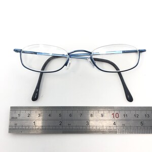 May include: Blue metal-framed eyeglasses with oval lenses and black temples. The glasses are positioned above a metal ruler, with the measurement scale visible. The frame has the text "46021 140" printed on the temple.