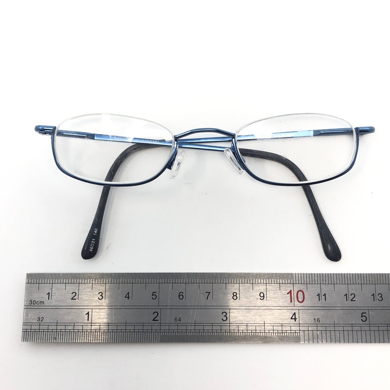 May include: Blue metal-framed eyeglasses with oval lenses and black temples. The glasses are positioned above a metal ruler, with the measurement scale visible. The frame has the text "46021 140" printed on the temple.