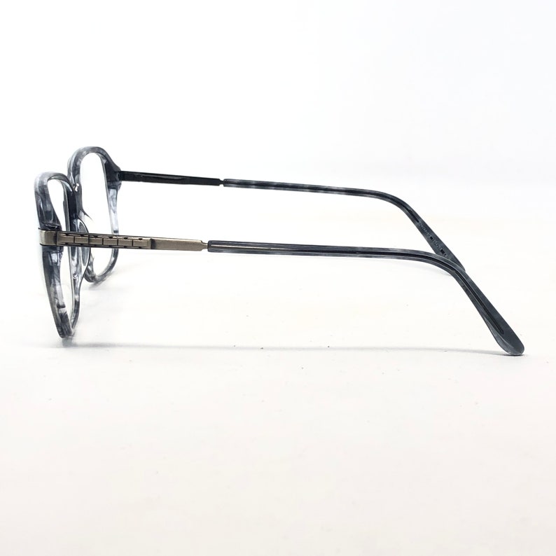 May include: Eyeglasses with a marbled grey and clear frame. The dark grey temples connect to the frame with a silver-coloured hinge. The clear lenses are set against a white background. The glasses are a classic style.