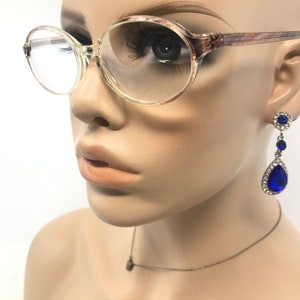 May include: Clear-framed spectacles with a brown and tan patterned design on the arms. The mannequin also wears blue teardrop earrings with a silver setting and a delicate silver necklace with a small pendant.