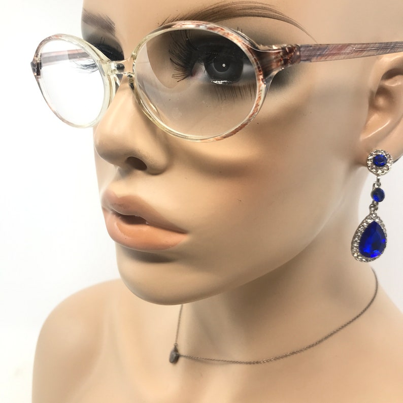 May include: Clear-framed spectacles with a brown and tan patterned design on the arms. The mannequin also wears blue teardrop earrings with a silver setting and a delicate silver necklace with a small pendant.