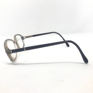 May include: A pair of eyeglasses with a clear frame and dark gray temples. The lenses are clear, and the frame has a rectangular blue accent on the side. The glasses are set against a white background.