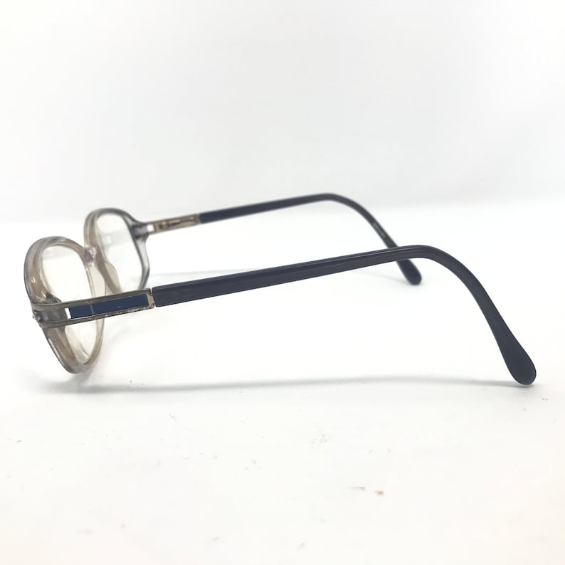 May include: A pair of eyeglasses with a clear frame and dark gray temples. The lenses are clear, and the frame has a rectangular blue accent on the side. The glasses are set against a white background.