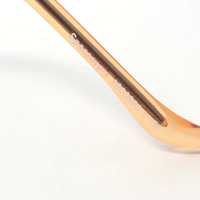 May include: Close-up of a gold-colored eyeglass frame arm. The text "Specsavers 1000880 L C" is printed on the arm. The frame is curved and has a smooth, metallic finish. The background is white.