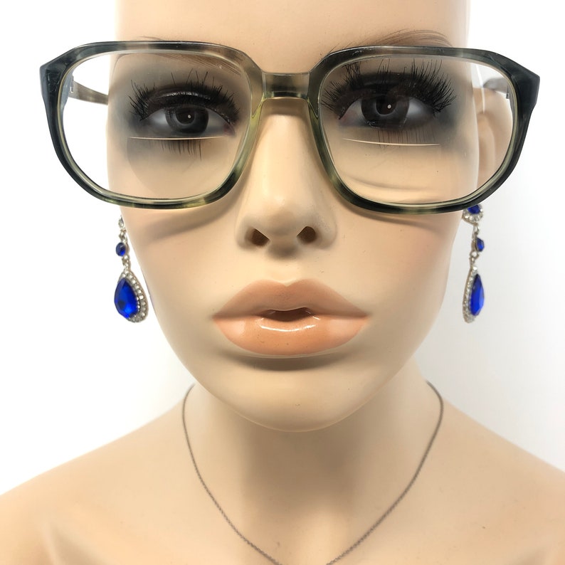 May include: A close-up of a mannequin wearing large, square-framed eyeglasses with a speckled green and black pattern. The mannequin also has long eyelashes and is wearing dangling earrings with blue teardrop-shaped gems.