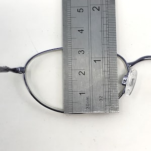 May include: Close-up of a pair of oval-shaped eyeglasses with a dark frame. A ruler is placed over the glasses, measuring approximately 1.5 inches across the lens. The glasses have clear lenses and nose pads.