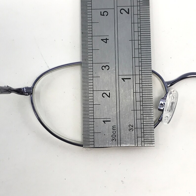 May include: Close-up of a pair of oval-shaped eyeglasses with a dark frame. A ruler is placed over the glasses, measuring approximately 1.5 inches across the lens. The glasses have clear lenses and nose pads.