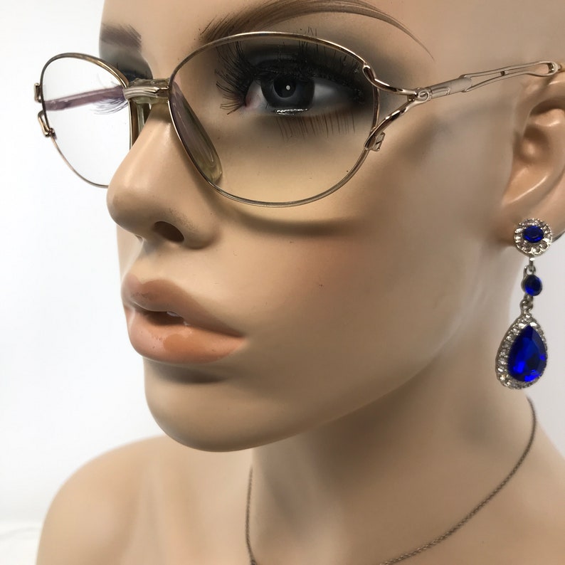 Peut inclure: Lunettes ovales dor&eacute;es avec des verres transparents. Les lunettes sont sur le visage d'un mannequin. Le mannequin porte des boucles d'oreilles bleues en forme de goutte d'eau avec des strass.