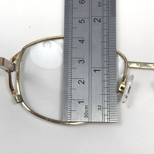 Op de afbeelding: Close-up van een goudomrande bril met heldere glazen. Een liniaal wordt naast de bril geplaatst en toont een afmeting van ongeveer 7,6 cm. De bril heeft een rechthoekige vorm en doorzichtige neuspads.