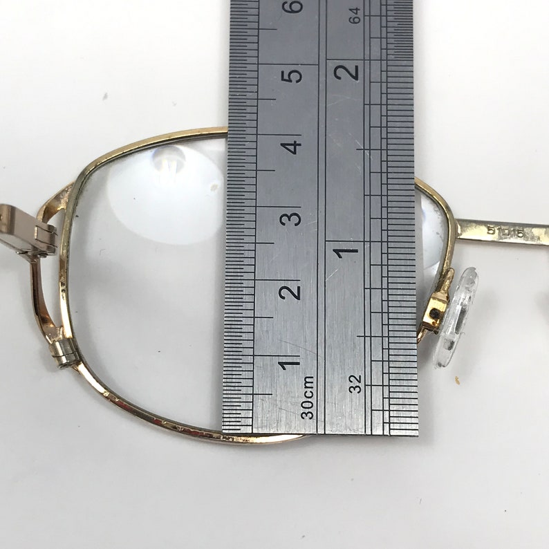 Op de afbeelding: Close-up van een goudomrande bril met heldere glazen. Een liniaal wordt naast de bril geplaatst en toont een afmeting van ongeveer 7,6 cm. De bril heeft een rechthoekige vorm en doorzichtige neuspads.