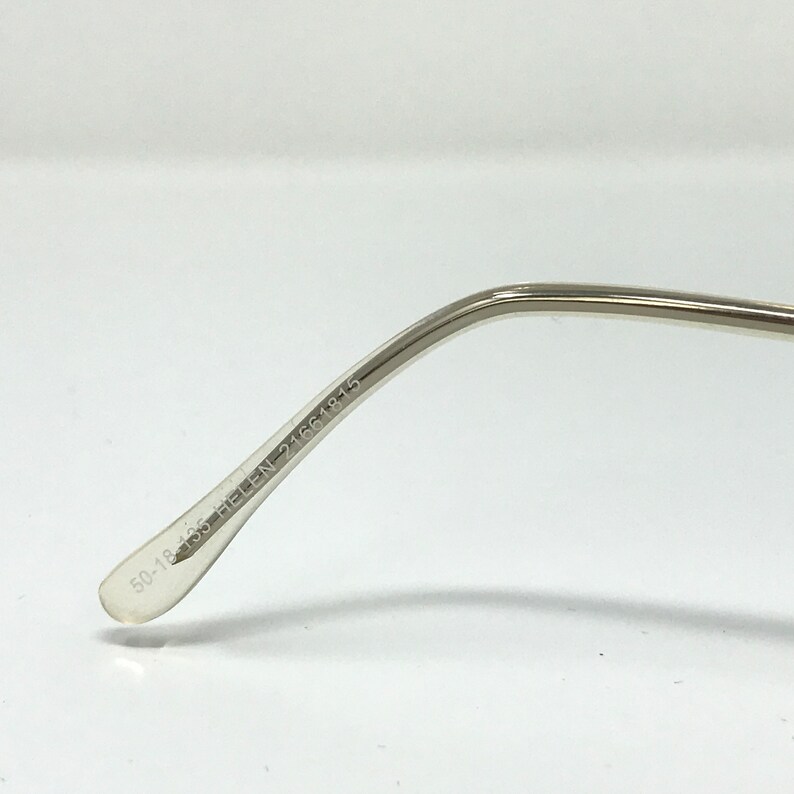 May include: Close-up of a silver-toned eyeglass frame arm. The text "50-18 135 HELEN-21661815" is printed on the inside of the arm. The frame has a clear, rounded tip.