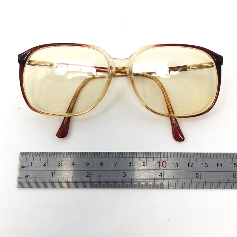 May include: Vintage eyeglasses with a brown and clear frame. The glasses have a rectangular shape with a gold-colored bridge and arms. A ruler is placed below the glasses, showing measurements in inches.
