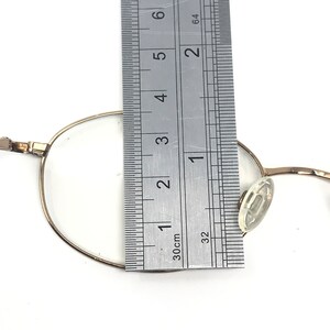 May include: A pair of gold-toned metal glasses with clear plastic earpieces. The glasses are measured against a ruler, showing the frame width is approximately 4.5 cm.