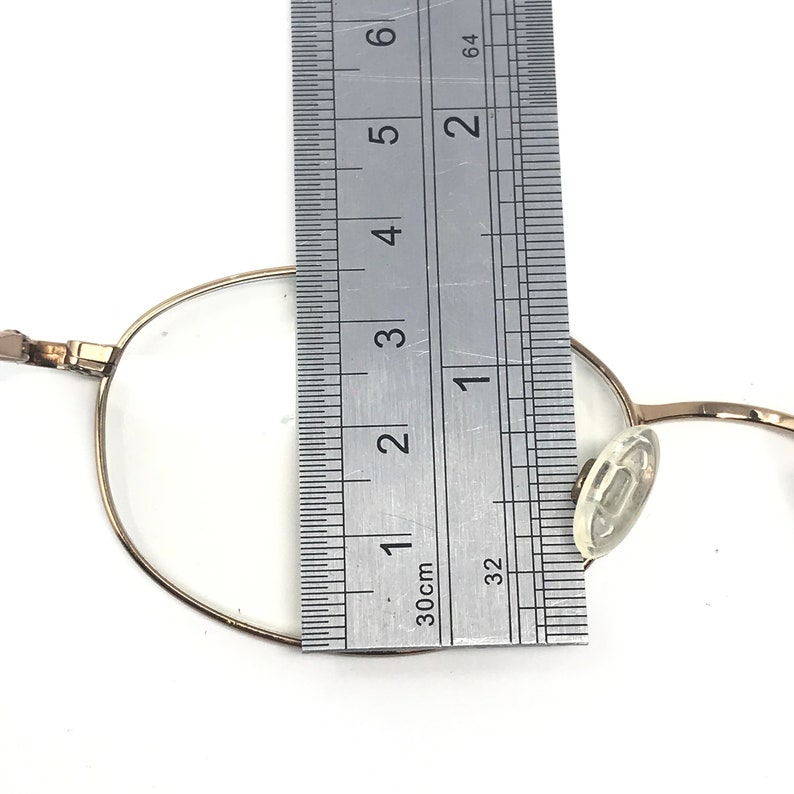 May include: A pair of gold-toned metal glasses with clear plastic earpieces. The glasses are measured against a ruler, showing the frame width is approximately 4.5 cm.