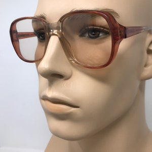 May include: Brown and clear square-framed eyeglasses. The frames have a gradient effect, transitioning from a darker brown to a lighter, almost transparent color. The lenses are clear, and the glasses are displayed on a mannequin.