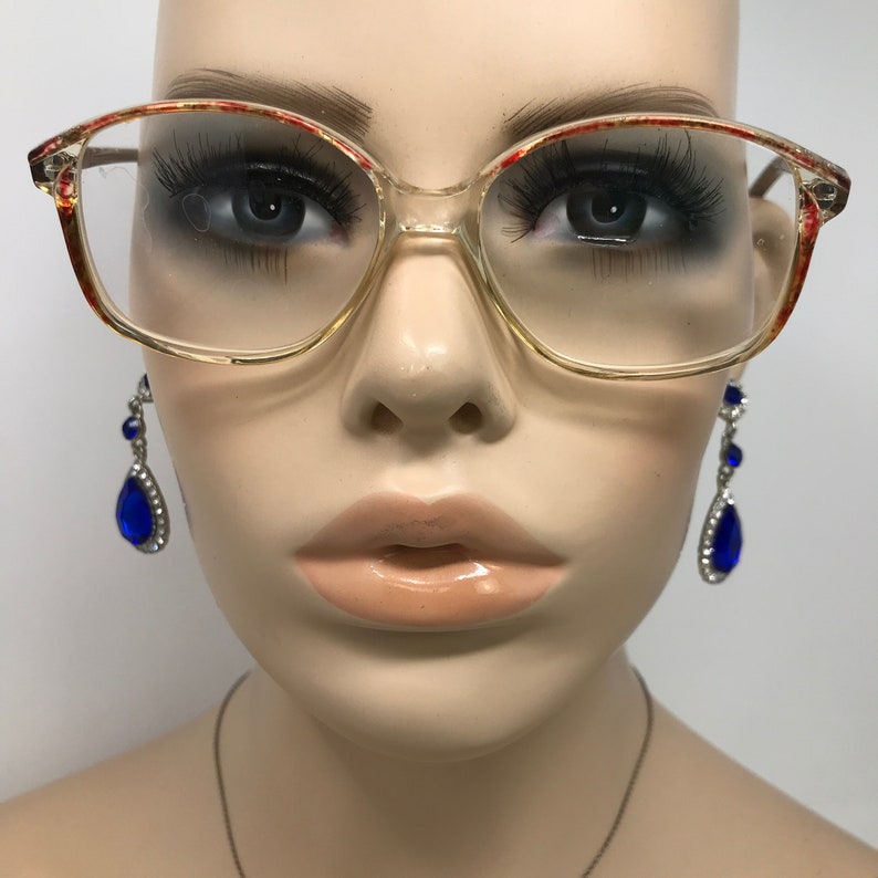 May include: Clear eyeglasses with a light brown frame and red accents on the upper corners. The glasses are on a mannequin head, with long eyelashes and blue teardrop earrings.