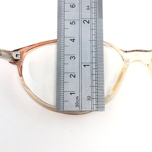 May include: Eyeglasses with a clear frame and a light brown upper frame. A ruler is placed over the glasses, measuring approximately 5 cm across the lens. The glasses are on a white background.
