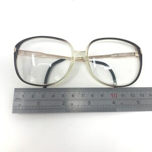 May include: Vintage eyeglasses with a clear frame and dark brown accents. The glasses have a rectangular shape and are measured with a ruler. The text "BRIAN 21001635" is visible on the frame.