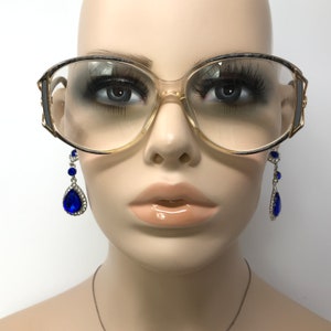 Puede incluir: Un par de gafas grandes, transparentes, con montura de pl&aacute;stico y detalles dorados. Las gafas est&aacute;n en una cabeza de maniqu&iacute; y el maniqu&iacute; lleva pendientes azules en forma de l&aacute;grima.