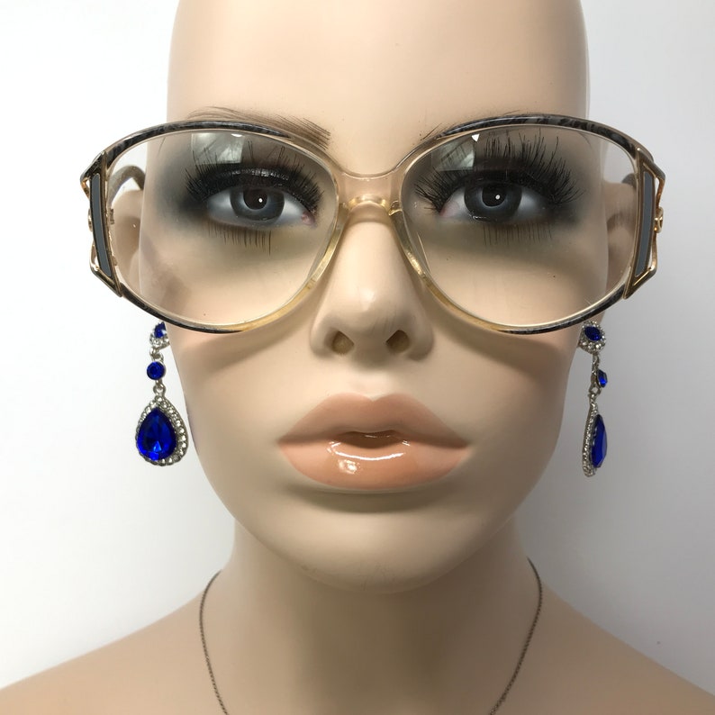 Puede incluir: Un par de gafas grandes, transparentes, con montura de pl&aacute;stico y detalles dorados. Las gafas est&aacute;n en una cabeza de maniqu&iacute; y el maniqu&iacute; lleva pendientes azules en forma de l&aacute;grima.