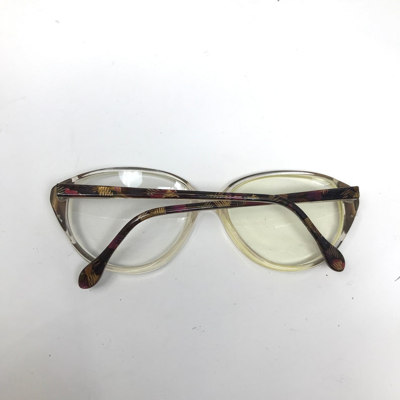 May include: Clear-framed eyeglasses with a patterned design on the temples and bridge. The frames have an oval shape. The temples feature a pattern of brown, yellow, and pink. The lenses are clear.