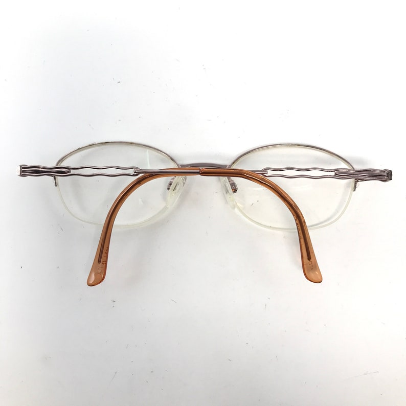 K&ouml;nnte beinhalten: Silberfarbene randlose Brille mit braunen B&uuml;geln. Die Brille hat ein zartes, wellenf&ouml;rmiges Design am Rahmen.