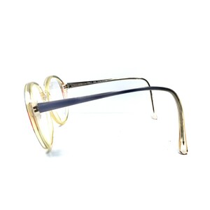 Puede incluir: Un par de gafas redondas, de pl&aacute;stico transparente, con patillas de metal azul. Las patillas est&aacute;n dobladas en un &aacute;ngulo de 90 grados y el texto "SPECSAVERS" est&aacute; impreso en la patilla derecha.