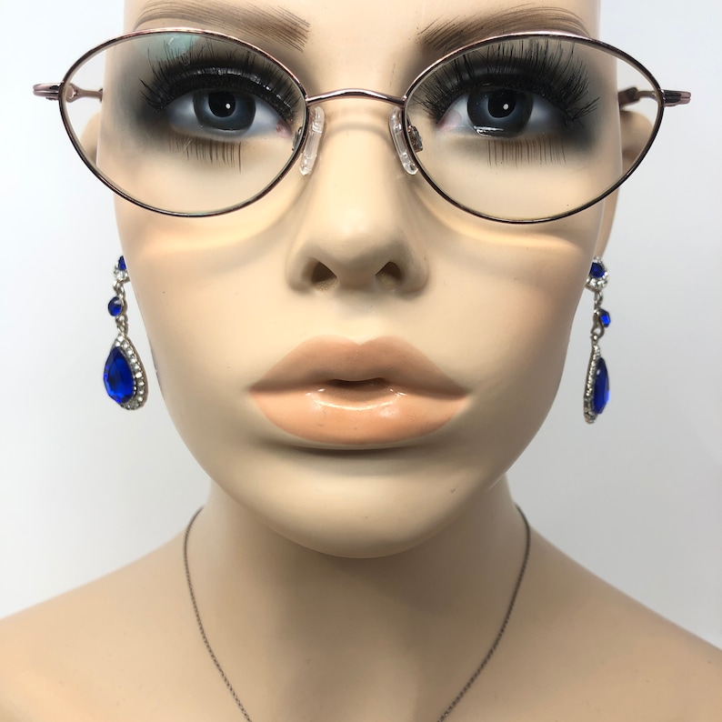K&ouml;nnte beinhalten: Ein Schaufensterpuppenkopf mit ovaler, ros&eacute;goldener Brille und baumelnden blauen Tr&auml;nenohrringen. Die Ohrringe sind mit klaren Kristallen verziert. Die Schaufensterpuppe hat lange Wimpern und eine neutrale Lippenfarbe.