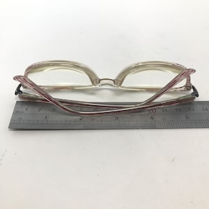 May include: Clear-framed eyeglasses with red and white striped temples. The glasses are positioned over a ruler, with the frame measuring approximately 5.5 inches in length. The lenses are clear, and the overall design is vintage.
