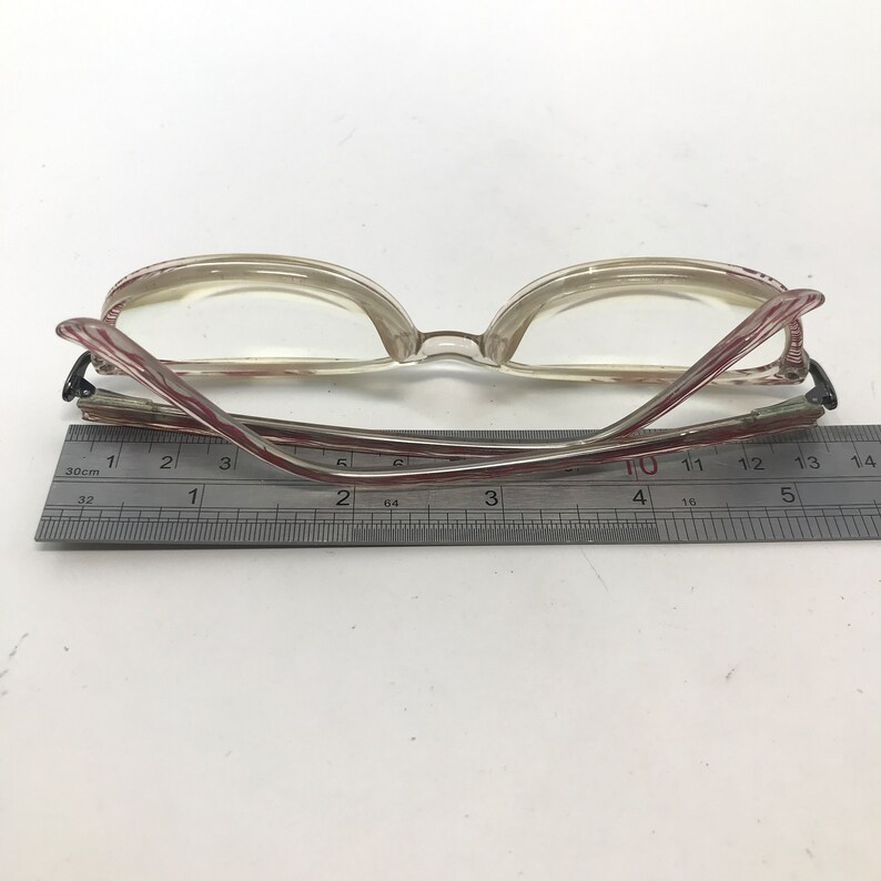 May include: Clear-framed eyeglasses with red and white striped temples. The glasses are positioned over a ruler, with the frame measuring approximately 5.5 inches in length. The lenses are clear, and the overall design is vintage.