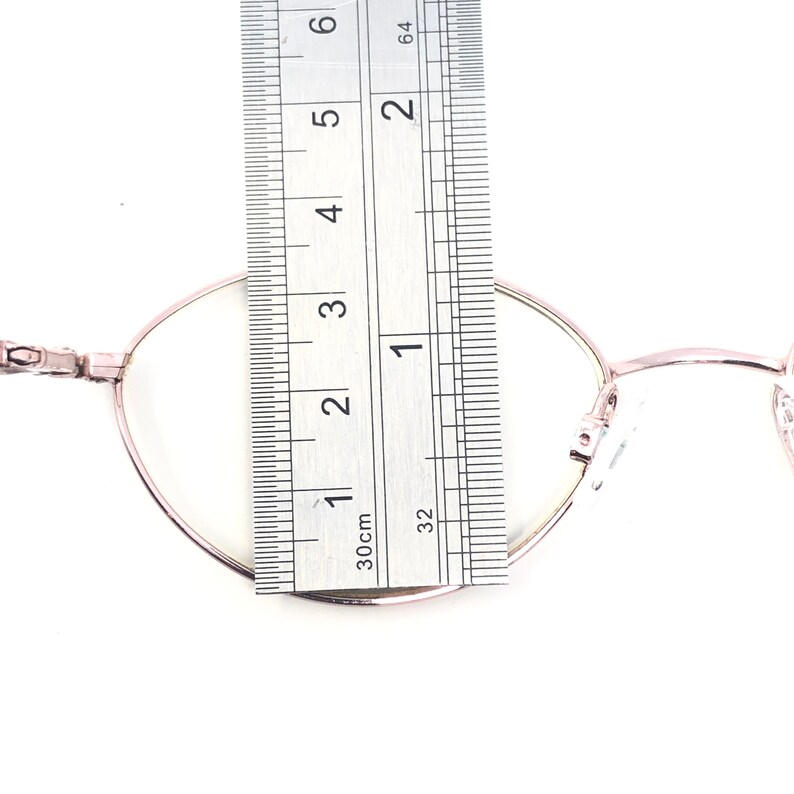 K&ouml;nnte beinhalten: Ros&eacute;goldene ovale Brille mit klaren Gl&auml;sern wird mit einem Lineal gemessen. Das Lineal zeigt, dass die Brille etwa 5 cm breit ist. Die Brille hat einen d&uuml;nnen Rahmen und ein klassisches Design.