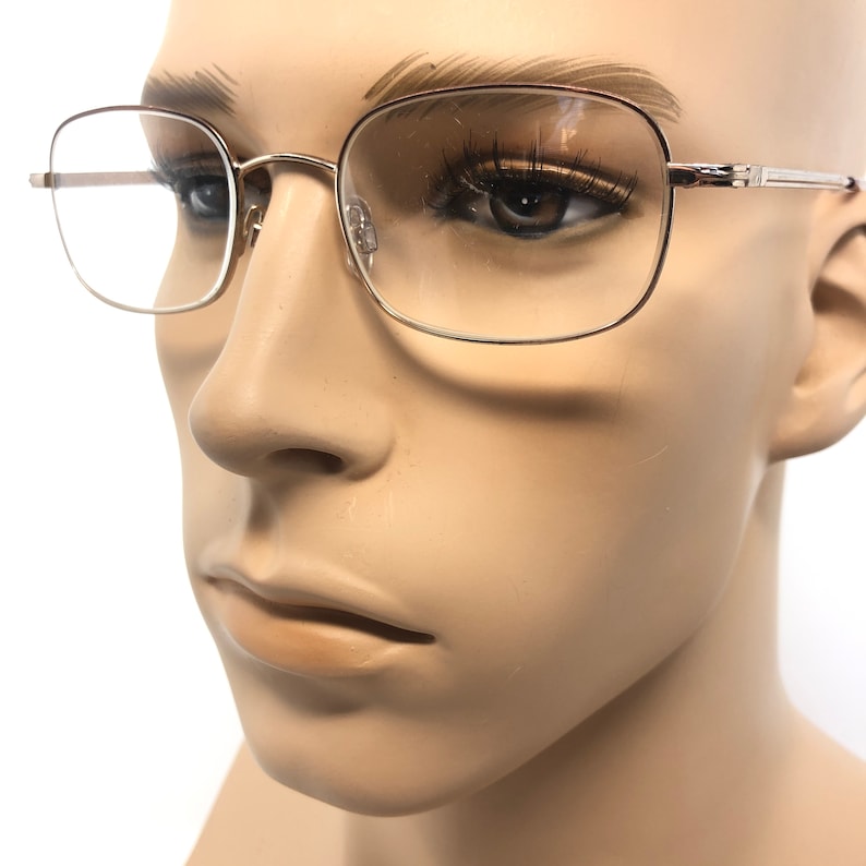 May include: Silver-framed spectacles with a rectangular shape. The glasses have clear lenses and are displayed on a mannequin. The frames are thin and have a classic design, suitable for everyday wear.