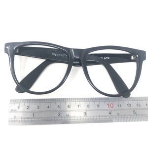 May include: Black eyeglasses with a thick frame, featuring the text "SPECSAV" and "BLACK". A ruler is placed below the glasses, showing measurements in inches. The glasses have a classic, rectangular shape.