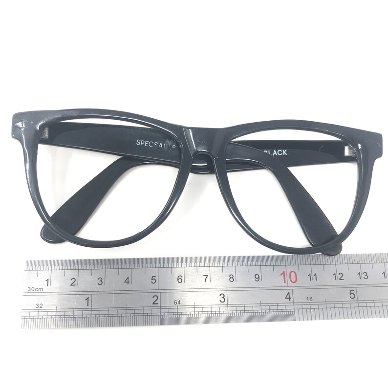 May include: Black eyeglasses with a thick frame, featuring the text "SPECSAV" and "BLACK". A ruler is placed below the glasses, showing measurements in inches. The glasses have a classic, rectangular shape.