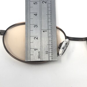 May include: Close-up of a pair of round eyeglasses with a dark frame and light-colored lenses. The glasses are being measured with a ruler, showing a length of approximately 1 inch.