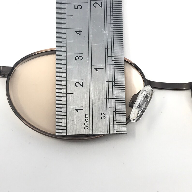May include: Close-up of a pair of round eyeglasses with a dark frame and light-colored lenses. The glasses are being measured with a ruler, showing a length of approximately 1 inch.