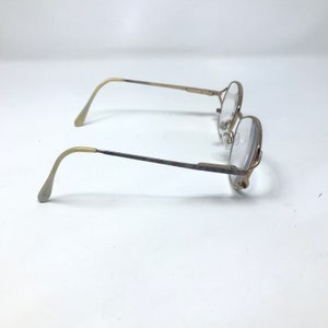 Op de afbeelding: Een vintage bril met een goudkleurig metalen montuur en heldere glazen. De bril heeft een klassieke ronde vorm en gebogen veren. De glazen zijn helder en het ontwerp is eenvoudig en elegant.