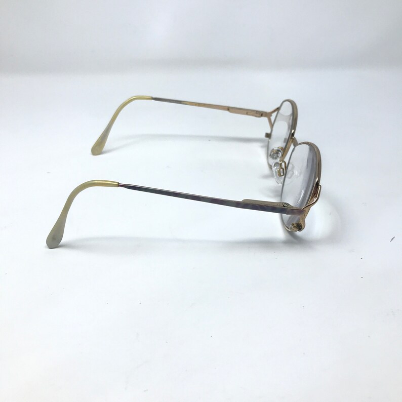 Op de afbeelding: Een vintage bril met een goudkleurig metalen montuur en heldere glazen. De bril heeft een klassieke ronde vorm en gebogen veren. De glazen zijn helder en het ontwerp is eenvoudig en elegant.